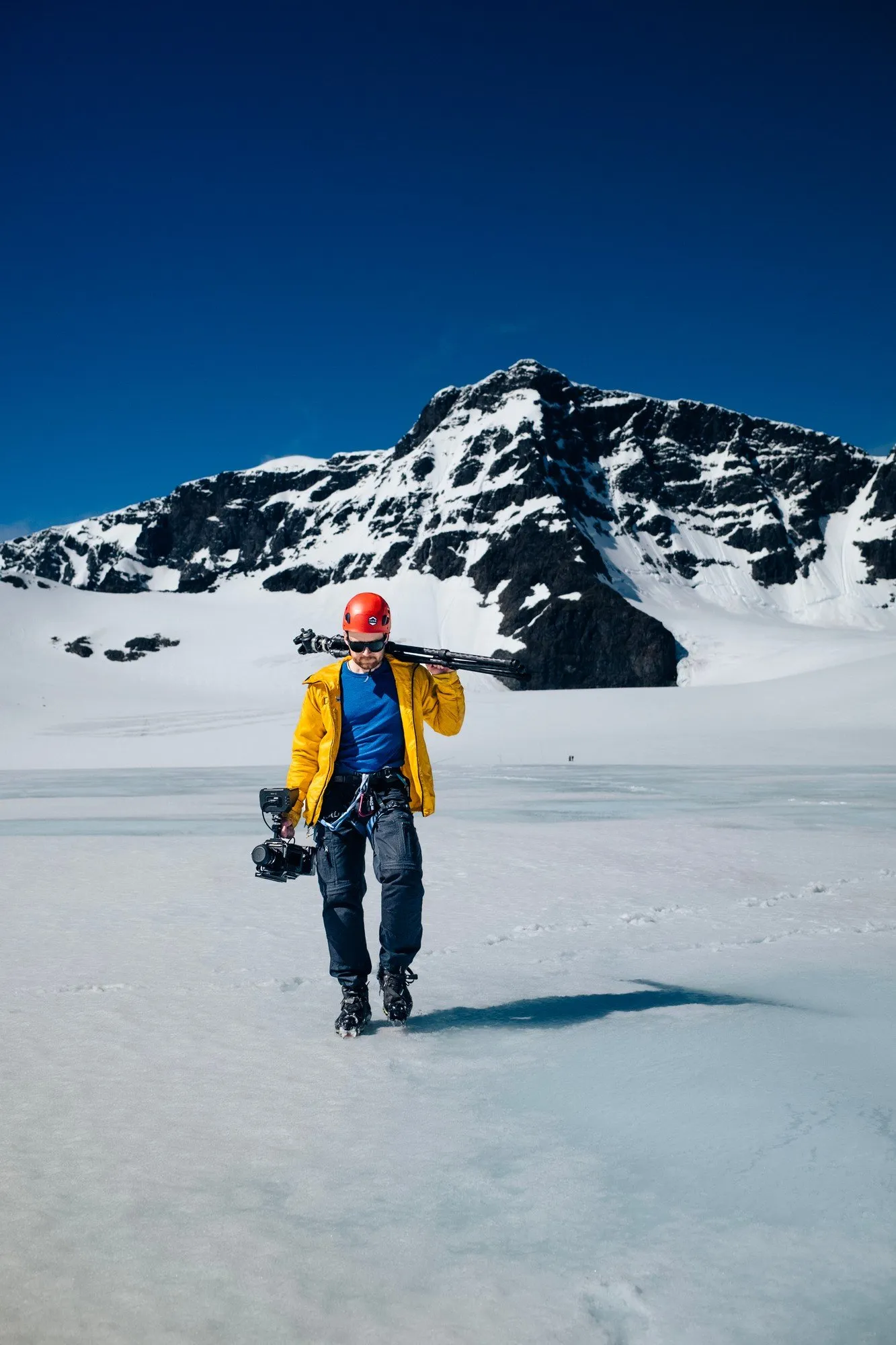 A crew member in a helmet carrying camera gear across Storglaciären under a blue sky
